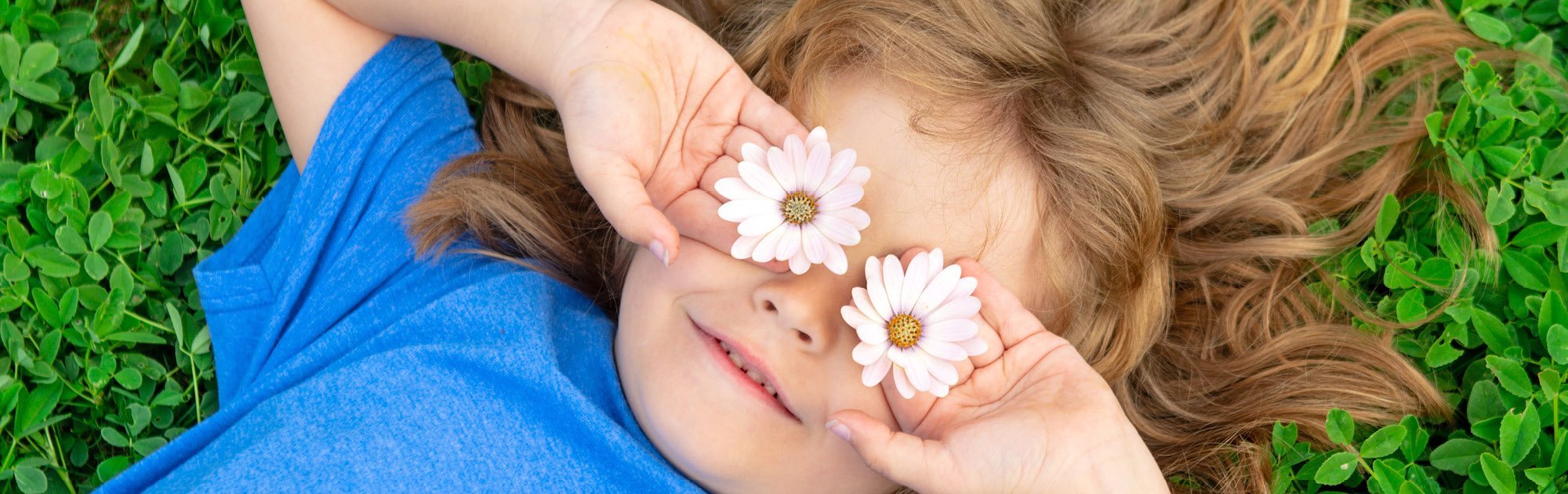 Kind liegt im Gras und hält zwei Gänseblümchen vor die Augen